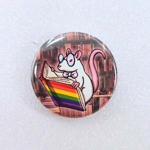 May include: A round button featuring a cartoon rat wearing glasses and a bow tie, reading a book with a rainbow-colored cover. The background shows a bookshelf. The rat has a pink tail.