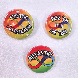 Könnte beinhalten: Drei runde, farbenfrohe Buttons mit Text und einem Unendlichkeitssymbol. Die Buttons haben einen Regenbogen-Farbverlauf im Hintergrund. Die Texte auf den Buttons lauten "EXISTING AUSTISTICALLY", "RIZZ 'EM WITH THE 'TISM" und "AUTASTIC!".