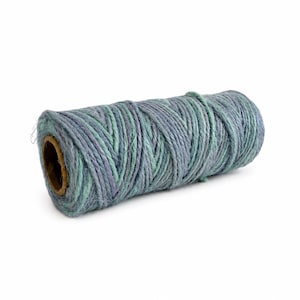 May include: A cylindrical spool of twisted twine featuring a variegated color palette of muted blue, soft purple, and light mint green fibers. The thick, textured cord is wound tightly around a central cardboard core, showing a multi-tonal appearance.