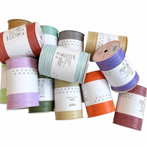 May include: A collection of twelve cylindrical spools of Ispie brand raffia yarn in various colors including red, light blue, lavender, orange, green, tan, and grey. Each spool features a white paper label with black text and a row of x symbols.