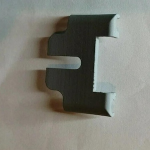 Puede incluir: Una pieza de plástico gris oscuro con una forma única, que presenta una abertura rectangular y múltiples protuberancias. Esta pieza es probablemente un componente de un ensamblaje más grande.