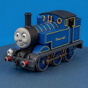 Peut inclure: Une locomotive à vapeur jouet bleue avec un visage gris souriant, une cheminée noire et des bordures dorées. Le côté de la machine porte l'inscription Thomas style en lettres dorées. Elle possède des roues bleues et un toit noir.