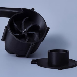 Può includere: Alloggiamento della girante nero stampato in 3D con un design a pale della ventola. L'alloggiamento è cilindrico con un ingresso curvo e diversi fori di montaggio. Sono visibili anche un componente cilindrico e una base separati.