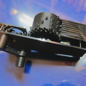 Peut inclure: Composant mécanique noir imprimé en 3D. Il présente une section cylindrique, des engrenages et un cadre rectangulaire avec des détails fendus. L'objet est posé sur un fond bleu. Le design est complexe.