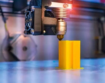 Puede incluir: Una impresora 3D extruye un objeto de plástico amarillo. La cabeza de impresión es visible en primer plano, con el objeto que se está imprimiendo en el fondo.