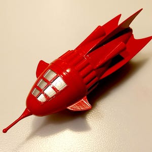 Puede incluir: Un cohete de juguete rojo brillante con una nariz puntiaguda y aletas. El cohete tiene una cabina con ventanas plateadas. El juguete probablemente está hecho de plástico o un material similar.