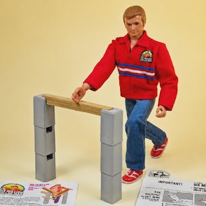 Op de afbeelding: Een mannelijk actiefiguur in een rood jasje met een Six Million Dollar Man-embleem, een blauwe spijkerbroek en rode sneakers. Het figuur staat naast een constructie van twee grijze kolommen van elk drie blokken, afgewerkt met een houten plank.