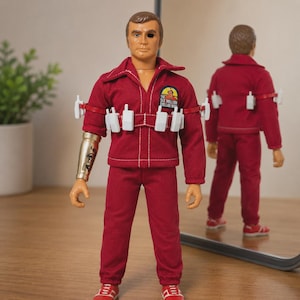 Op de afbeelding: Een Six Million Dollar Man actiefiguur in een rode jumpsuit met een witte riem waaraan verschillende witte walkietalkie-accessoires zijn bevestigd. Het figuur heeft een metalen rechterarm, een zwarte ooglap en een logo op de borst.