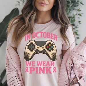 Puede incluir: Camiseta rosa claro con el texto "IN OCTOBER WE WEAR PINK" y un gráfico de un mando de videojuegos. El mando es beige y gris con botones de colores. También presenta lazos rosas.
