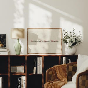 Puede incluir: Un cuadro enmarcado con el texto "all first drafts of everything are garbage" en rojo y negro. El cuadro se exhibe sobre una estanter&iacute;a de madera oscura con libros, una radio y una l&aacute;mpara verde. Un jarr&oacute;n con flores blancas est&aacute; cerca.