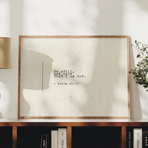 Puede incluir: Impresión enmarcada con el texto "Be still, and know that I am God. - Salmo 46:10". La impresión tiene un fondo beige claro y un marco de madera. La obra de arte se exhibe en una estantería.