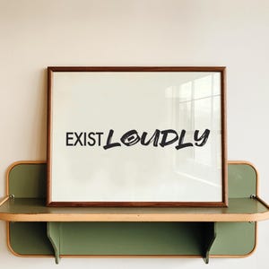 May include: A rectangular wall art print in a dark brown wooden frame featuring the black text "EXIST LOUDLY" in a bold, brush-stroke style font. The print is set against a light off-white background, creating a high-contrast visual display.