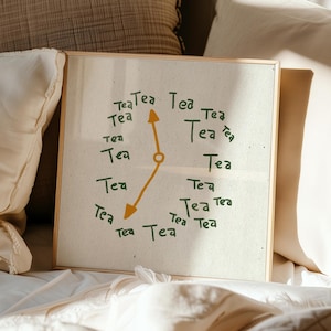 Könnte beinhalten: Ein quadratisches Whiteboard mit goldenem Rahmen zeigt ein Uhrdesign. Das Zifferblatt zeigt das Wort "Tea" in Grün anstelle von Zahlen. Orangefarbene Zeiger zeigen die Uhrzeit an. Das Whiteboard liegt auf einem Bett.