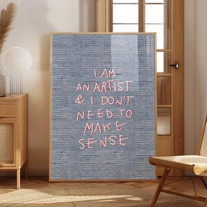 以下が含まれることがあります： 「I AM AN ARTIST & I DON'T NEED TO MAKE SENSE」の赤い文字が、テクスチャ加工されたグレーの背景に描かれた額入りのアートプリント。 フレームは明るい茶色です。