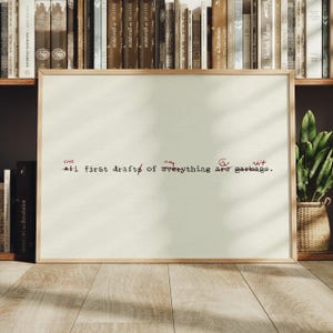 May include: A rectangular framed print with a cream-colored background featuring black typewriter-style text that reads: the any is shit All first drafts of everything are garbage. The words are crossed out with red ink marks and handwritten red edits.
