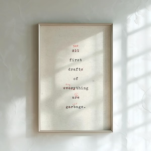 Puede incluir: Obra de arte enmarcada con el texto "The All first drafts of any everything are shit garbage" en fuente de máquina de escribir roja y negra. La obra de arte está en un marco plateado y cuelga de una pared blanca.