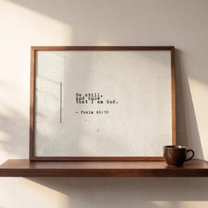 May include: A rectangular wooden frame containing a white textured print with the black typewriter-style text: Be still, and know that I am God. - Psalm 46:10. A vertical black line appears to the left of the text on the off-white background.