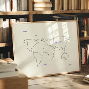 Pode incluir: Uma impressão emoldurada com um contorno preto minimalista do mapa-múndi sobre um fundo creme. O texto azul diz "Home is wherever you are". A obra de arte é apresentada numa moldura de madeira, ideal para decoração de interiores.