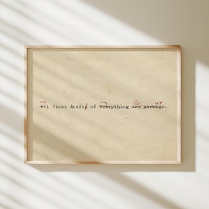 May include: A framed print with the text "All first drafts of everything are garbage." The text is in black typewriter font with red handwritten additions. The paper has a vintage, aged appearance, and the frame is light wood.