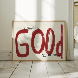 May include: A rectangular wall art print in a light wood frame featuring the red hand-lettered text IT FEELS SO GOOD TO BE HOME on a textured off-white background. The word GOOD is written in large, bold red letters with a brushstroke appearance.