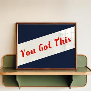 May include: A rectangular framed print featuring a dark blue background with a wide, diagonal white stripe running from the bottom left to the top right. The text "You Got This" is printed in a bold, red, cursive font across the white stripe.
