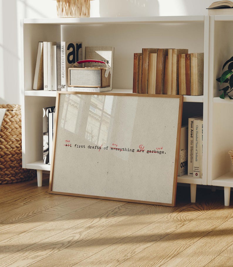 Puede incluir: Un cuadro enmarcado con el texto "+1 first drafts of everything are garbage" en rojo y negro. El cuadro se exhibe sobre un suelo de madera frente a una estanter&iacute;a blanca llena de libros y una peque&ntilde;a radio.