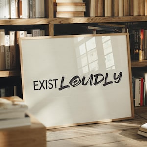 May include: A rectangular framed print with a light beige background and the black text "EXIST LOUDLY" written in a bold, brush-stroke style font. The frame is made of light-coloured wood and is positioned in front of a wooden bookshelf.