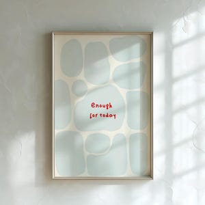 May include: A framed vertical wall art print featuring a cream-colored background with scattered, soft light blue organic shapes. Centered in the middle of the composition, the text "Enough for today" is written in a simple, bold red font.