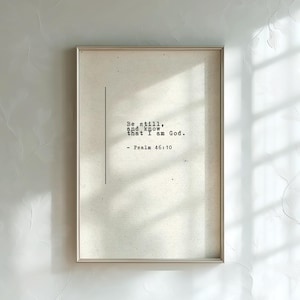 Puede incluir: Un cuadro rectangular con marco que presenta un fondo de color beige claro texturizado. El texto "Be still, and know that I am God. - Psalm 46:10" aparece en una fuente negra estilo máquina de escribir, junto a una línea vertical negra.