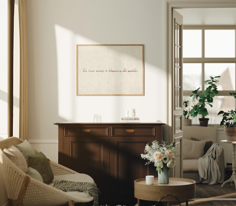 Puede incluir: Un cuadro enmarcado con el texto "Art. First draft of everything are garbage." cuelga sobre un mueble de madera marr&oacute;n oscuro. Una silla de mimbre y una mesa de centro redonda de madera con un jarr&oacute;n de flores est&aacute;n en primer plano.
