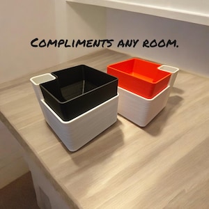 Peut inclure: Deux contenants de rangement carrés empilés avec une couche extérieure blanche et une couche intérieure noire et orange. Le texte "COMPLIMENTS ANY ROOM." est écrit en noir sur une surface blanche.