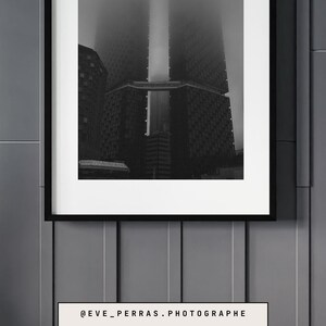 Peut inclure: Photographie en noir et blanc d'un immeuble haut dans le brouillard. Le bâtiment est en béton et présente un design géométrique. Le brouillard est épais et obscurcit le sommet du bâtiment. L'image est encadrée dans un cadre noir.