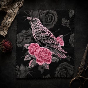 May include: A rectangular fabric patch featuring a black background with a tonal rose pattern. Centered is an embroidered design of a crow in shades of pink and grey, perched atop two vibrant pink roses with green leaves and grey accents.