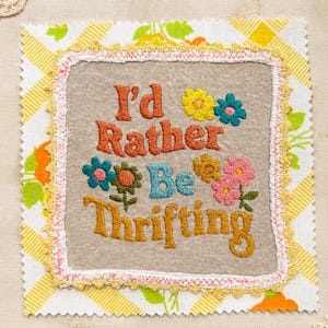 May include: A square fabric patch featuring the embroidered text I'd Rather Be Thrifting in orange, blue, and yellow thread. The design includes colorful embroidered flowers and is bordered by a delicate pink and yellow crocheted lace trim.