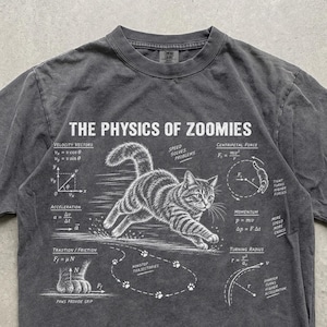Op de afbeelding: Een antracietgrijs t-shirt met korte mouwen en een witte afbeelding van een rennende kat met de tekst THE PHYSICS OF ZOOMIES. Het ontwerp bevat diverse natuurkundige vergelijkingen, diagrammen en labels zoals Velocity Vectors, Acceleration, Traction, Friction, Centripetal Force, Momentum en Turning Radius.