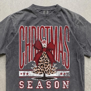 Może przedstawiać: Ciemnoszary t-shirt z krótkim rękawem z czerwonym napisem CHRISTMAS SEASON. Centralna grafika przedstawia stylizowaną choinkę w panterkę, zwieńczoną dużą czerwoną kokardą i małą gwiazdką, umieszczoną na tkaninie o efekcie sprania.