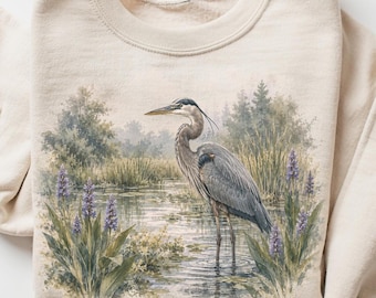 May include: A sand-colored crewneck sweatshirt featuring a detailed graphic print of a great blue heron standing in a marshy wetland environment with green reeds, tall grasses, and purple wildflowers. The text SAND appears in a small circle.