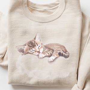 May include: A sand-coloured sweatshirt with a graphic of a sleeping kitten on a cloud. The kitten is light brown with darker stripes. The sweatshirt has a crew neck and long sleeves, with the right sleeve cuffed. The word "SAND" is printed on a small circle.
