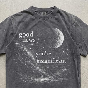 May include: A charcoal gray short-sleeve t-shirt featuring a white graphic of a moon, stars, and a small figure standing on a landscape. The text reads "good news" and "you're insignificant" in a white serif font. The shirt has a crew neckline.