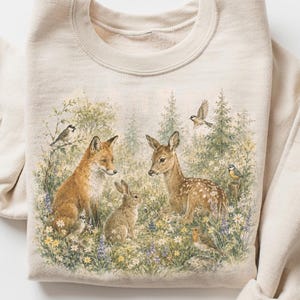 May include: A sand-colored crewneck sweatshirt featuring a detailed graphic print of a forest scene. The illustration depicts a fox, a rabbit, and a fawn surrounded by various small birds, green trees, and colorful wildflowers in a meadow setting.