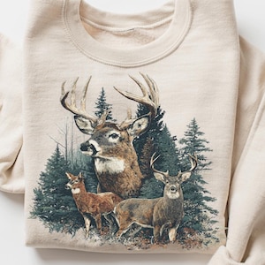 Op de afbeelding: Een zandkleurige sweatshirt met een gedetailleerde illustratie van drie herten in een bosrijke omgeving. Het ontwerp omvat een grote bok met prominente geweien, twee kleinere herten en groenblijvende bomen. Het woord "SAND" is gedrukt op een klein, rond label.