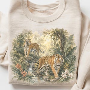 Op de afbeelding: Zandkleurige sweatshirt met een gedetailleerde illustratie van twee tijgers in een weelderige jungle. De tijgers hebben oranje en zwarte strepen, omringd door groene bladeren, bloemen en een waterval. De sweatshirt heeft een ronde hals en lange mouwen.