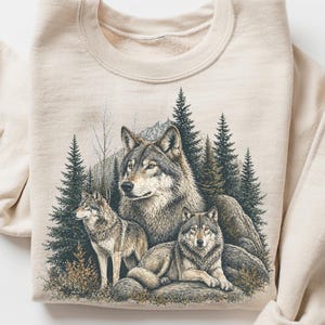 May include: A sand-coloured sweatshirt with a detailed illustration of three wolves in a forest setting. The design includes evergreen trees, rocks, and a mountain backdrop. The word "SAND" is printed on a small circle.