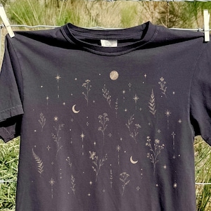 Könnte beinhalten: Ein schwarzes Kurzarm-T-Shirt mit einem Aufdruck aus verschiedenen Wildblumen, Farnen, Mondsicheln und kleinen Sternen in hellem Beige. Das Motiv ist mittig auf der Vorderseite des T-Shirts platziert, das über einen klassischen Rundhalsausschnitt verfügt.