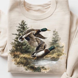 Puede incluir: Sudadera color arena con una ilustración detallada de dos patos ánades reales en vuelo sobre el agua. Los patos tienen cabezas verdes, alas marrones y cuerpos blancos y marrones. El fondo incluye árboles y follaje.