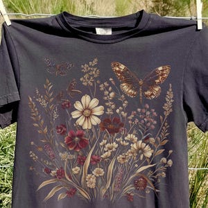May include: A charcoal black short-sleeve t-shirt featuring a central graphic print of a wildflower bouquet in muted earth tones, including cream, rust, and brown flowers, accompanied by two detailed butterflies hovering above the floral arrangement.