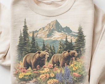 May include: Sand-colored sweatshirt featuring a scenic illustration of two brown bears in a meadow with colorful wildflowers, pine trees, and a mountain range. The sweatshirt has a crew neck and long sleeves.