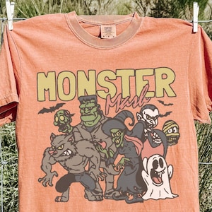 May include: A faded orange short-sleeve t-shirt featuring a graphic print with the text "MONSTER Mash" in yellow and red. The illustration depicts a group of cartoon monsters including a werewolf, Frankenstein's monster, a witch, a vampire, a ghost, and a mummy.
