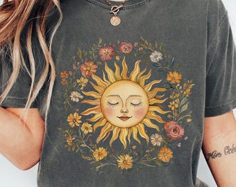 May include: Dark grey t-shirt featuring a sun graphic with closed eyes, surrounded by a floral wreath. The sun is yellow and orange, and the flowers are various colors. The shirt has a relaxed fit.