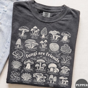 Puede incluir: Una camiseta gris carbón con un estampado gráfico blanco de varias especies de hongos con sus nombres etiquetados debajo de cada uno, y el texto "fungi are friends" en el centro. La camiseta tiene cuello redondo y una etiqueta Comfort Colors.